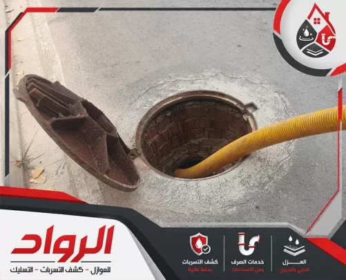 فتحة صرف أرضية مفتوحة في الطريق، مع إدخال خرطوم شفط أصفر داخلها لسحب المياه أو المخلفات المتراكمة.